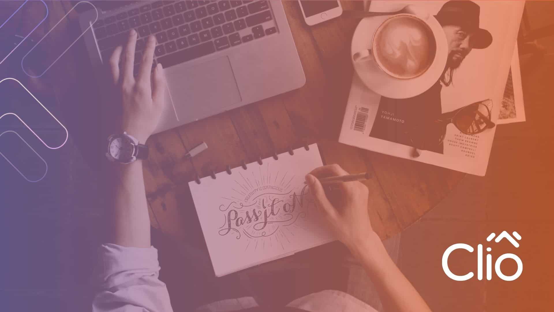 A person sketches "Pass It On" in a notebook at a wooden desk with a laptop, coffee, magazine, and phone—subtly inspired by 8 BUSINESS BLOGGING TACTICS TO GENERATE MORE SALES. The Clio logo glows in the lower right; a warm, gradient overlay brightens the scene. - Clio Websites | Top Canadian Web Design Company